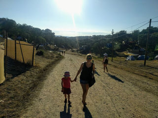 Dusty sunlit path winding through a vibrant forest campsite at Boom Festival, Portugal; families and friends stroll, soaking up the laid-back, communal atmosphere after years apart.