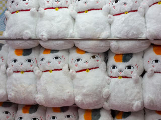 Japanese souvenir stall display, rows of fluffy white maneki-neko plushies with red collars, golden bells, and playful calico ears—a cheerful Kyushu travel keepsake.