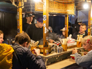Crowded Fukuoka yatai ramen stall, steaming bowls, bamboo chopsticks, handwritten Japanese menus, warm lantern light, travelers and locals shoulder-to-shoulder.