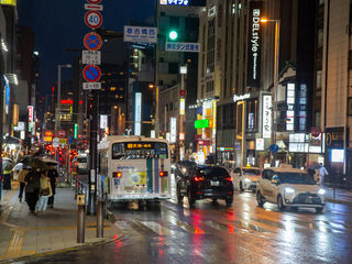 Rainy-night Fukuoka at Haruyoshi-bashi-nishi: neon-lit streets, Nishitetsu buses, umbrellas, taxis, glowing signs, and rain-slick pavement—Kyushu’s lively urban rhythm.