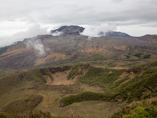Mount Aso’s vast volcanic caldera, rugged ash-brown slopes, emerald grasslands, winding roads, and a shifting cloud veil across Kyushu’s highland landscape.