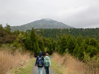 Misty Kyushu mountain landscape; volcanic slopes beneath low clouds, dense cedar forests, tawny grasses, and two hikers along a quiet trail.
