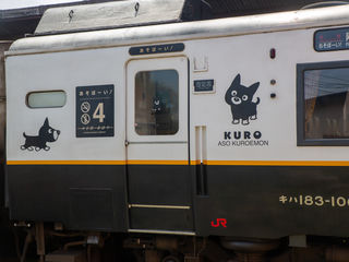 Kyushu Railway’s Aso Boy! carriage, featuring playful Kuroemon artwork, Japanese signage, reserved seating, and weathered black-and-white styling at Aso Station.