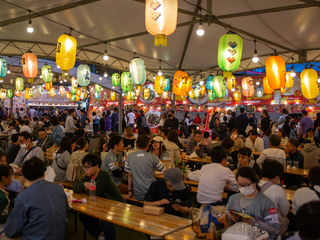 Fukuoka festival food hall, glowing paper lanterns, steaming ramen stalls, crowded wooden tables, Japanese street-food aromas, convivial evening energy beneath a tented canopy.