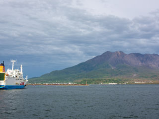 Kagoshima’s Sakurajima above Kagoshima Bay; volcanic slopes, layered clouds, a brightly painted ferry, and dark, wind-ruffled water.
