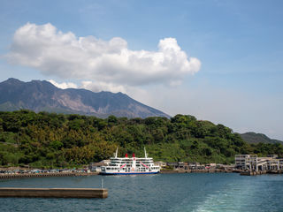 Kagoshima’s Sakurajima Ferry beneath volcanic Mount Sakurajima, with lush subtropical hills, sparkling harbor water, and dramatic clouds over southern Kyushu.